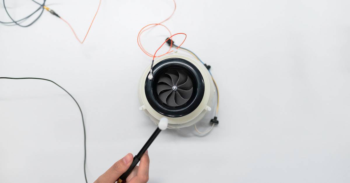 A hand holding a black screwdriver, adjusting a small jet engine model with visible wires on a white background to ensure product safety.